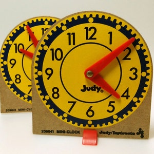 May include: Two vintage Judy Mini-Clocks with yellow faces, black numerals, and red hands. The clock faces have a blue gear-like border and the word "Judy" printed on them. The clocks are set at approximately 10:10. The base is made of cardboard.
