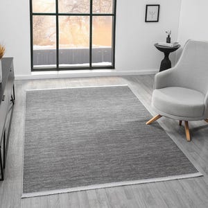 May include: A rectangular area rug featuring a heathered charcoal grey texture with subtle horizontal variations in tone. The rug has a clean, straight-edged border and a short, light-coloured fringe detail along the shorter ends of the piece.