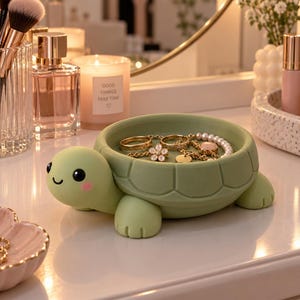 Pode incluir: Um porta-joias em forma de tartaruga verde-claro com acabamento mate liso. A tartaruga tem um rosto sorridente com olhos pretos e bochechas rosadas. O casco serve como uma taça contendo anéis dourados, um pingente de flor e uma pulseira de pérolas.