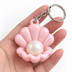 May include: A light pink seashell-shaped keychain featuring a smooth, glossy finish. The shell is open, revealing a large, round, iridescent white pearl centered within the shell. A silver-toned metal split ring is attached to the top loop.