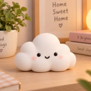 Puede incluir: Una luz nocturna con forma de nube blanca y acabado mate suave. La lámpara presenta un dibujo lineal sencillo en negro de una cara sonriente con dos pequeños ojos circulares negros, una boca curvada y mejillas circulares de color rosa suave.
