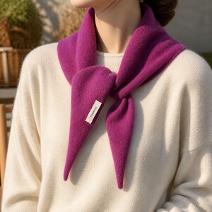 May include: A vibrant magenta knit scarf tied around the neck, featuring a soft, textured fabric and a small white rectangular label with the black text YOI&WOOL stitched onto one of the pointed ends, worn over a cream-coloured long-sleeve jumper.