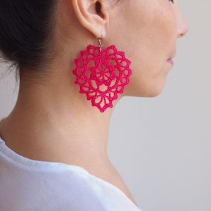 Op de afbeelding: Een paar roze gecrochete hartvormige oorbellen. De oorbellen zijn delicaat en ingewikkeld, met een bloemmotief.