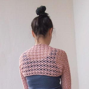 May include: A light pink, open-weave crochet shrug with long sleeves. The shrug is worn over a dark blue top. The crochet pattern creates a delicate, airy texture. The background is a plain white wall.