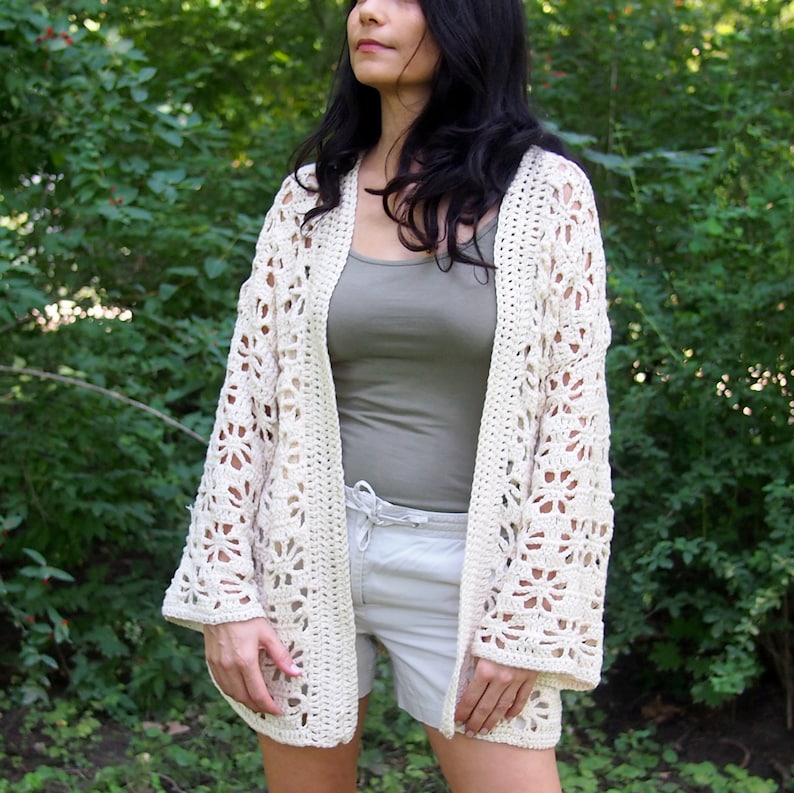 K&ouml;nnte beinhalten: Ein wei&szlig;er H&auml;kelcardigan mit einem Spitzenmuster. Der Cardigan ist vorne offen und hat lange &Auml;rmel. Das Model tr&auml;gt den Cardigan &uuml;ber einem grauen Tanktop und einer wei&szlig;en Shorts.