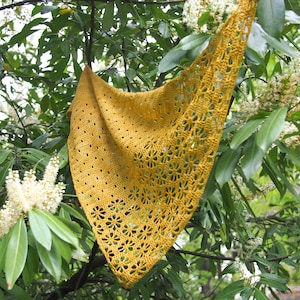 Op de afbeelding: Een gele gehaakte sjaal met een delicaat bloemenpatroon hangt aan een tak. De sjaal is gemaakt van fijn garen en heeft een zacht, luchtig gevoel.