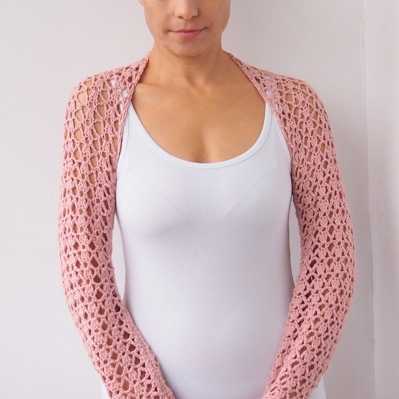 May include: A light pink, open-weave crochet shrug with long sleeves. The shrug is worn over a white tank top. The crochet pattern creates a delicate, airy texture. The garment is designed to be worn over the shoulders and arms.