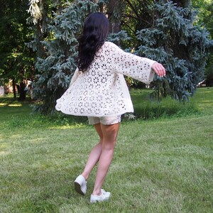K&ouml;nnte beinhalten: Ein wei&szlig;er H&auml;kelcardigan mit Blumenmuster. Der Cardigan ist vorne offen und hat lange &Auml;rmel. Die Person, die den Cardigan tr&auml;gt, steht in einem grasbewachsenen Bereich.