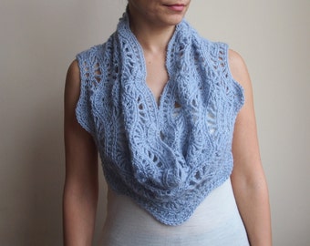May include: A light blue, sleeveless crochet cowl scarf. The openwork design features a repeating pattern of vertical lines and floral-like shapes. The cowl is worn around the neck and shoulders, creating a layered look. The soft blue color adds a touch of elegance.