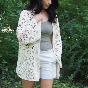 K&ouml;nnte beinhalten: Ein wei&szlig;er H&auml;kelcardigan mit einem Spitzenmuster. Der Cardigan ist vorne offen und hat lange &Auml;rmel.