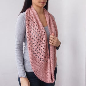 May include: A dusty rose crochet infinity scarf. The scarf features an open, lacy design with a solid border. The soft, lightweight scarf is worn around the neck and shoulders. This handmade accessory is perfect for adding a touch of elegance to any outfit.
