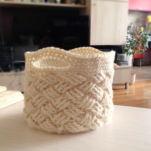 May include: A white crocheted basket with a braided pattern. The basket has a wide, rounded opening and a handle.