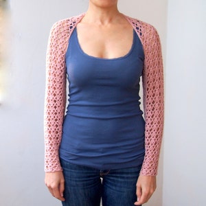 May include: A light pink, open-weave crochet shrug with long sleeves. The shrug is worn over a blue tank top and jeans. The crochet design features a repeating pattern of open spaces, creating a delicate and airy look.