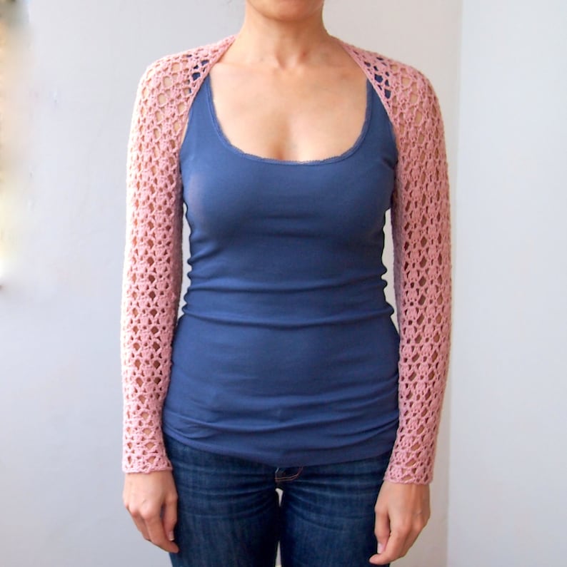 May include: A light pink, open-weave crochet shrug with long sleeves. The shrug is worn over a blue tank top and jeans. The crochet design features a repeating pattern of open spaces, creating a delicate and airy look.