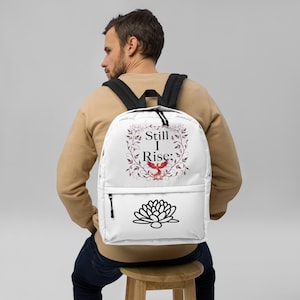 May include: A white backpack featuring black straps and a front graphic. The design includes the text Still I Rise; surrounded by a floral wreath and a red phoenix illustration. Below this, a black line art lotus flower is printed on the pocket.
