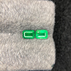 May include: Two vibrant green emerald gemstones with a rectangular step-cut facet pattern. The stones are positioned side-by-side, displaying a clear, transparent quality and a deep, saturated green hue characteristic of natural emeralds.