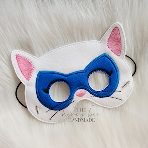 May include: White felt cat mask with blue eye mask and pink ears. The mask has black embroidered whiskers and the text "THE honey bee HANDMADE" is printed on the bottom.