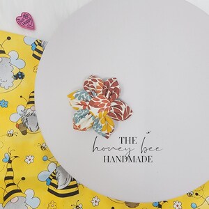 May include: A fabric flower with patterned petals in brown, orange, yellow, and blue. The flower is on a white surface with the text "THE honey bee HANDMADE". A yellow fabric with gnomes and bees is in the background.