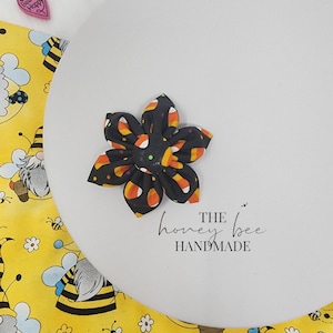 May include: A black fabric flower with a candy corn print. The flower is handmade and has a black centre with green and orange polka dots. The text "THE honey bee HANDMADE" is printed below the flower.