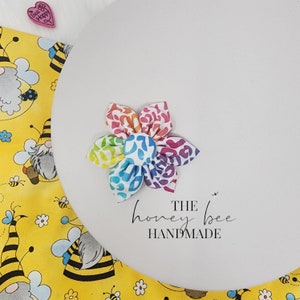 May include: A rainbow-coloured fabric flower with a white centre. The flower is made of a soft, textured fabric and has a slightly ruffled appearance. The flower is on a white background with the text "THE honey bee HANDMADE" below it.