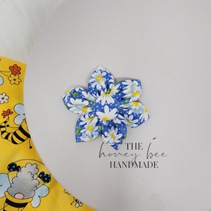 May include: A blue and white fabric flower with a daisy print. The flower is handmade and has a centre button. The text "THE honey bee HANDMADE" is below the flower.
