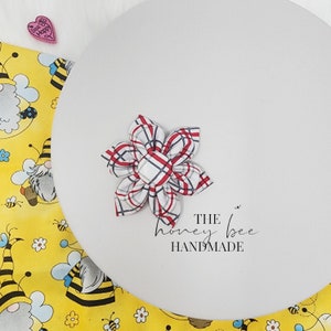 May include: A red and white plaid fabric flower hair clip with a white button centre. The clip is on a white background with the text "THE honey bee HANDMADE".