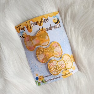 May include: Three yellow and beige fabric hair bows with gold accents. The bows are on a white card with the text "The Honeybee Handmade" and a bee illustration.