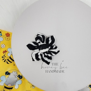 May include: Black and white zebra patterned fabric flower hair bow. The bow is handmade and has a centre made of the same fabric. The text "THE honey bee HANDMADE" is printed on a white background.