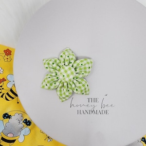 May include: A green and white gingham fabric flower with a white daisy print. The flower is handmade and has a ruffled appearance. The text "THE honey bee HANDMADE" is visible below the flower.