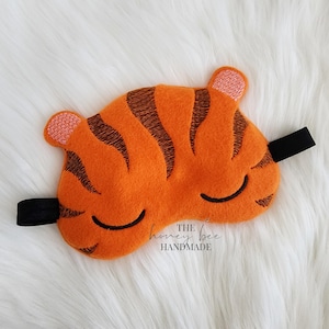 May include: Orange sleep mask with black stripes and a black elastic band. The mask is shaped like a tiger's face, with closed eyes and small ears. The text "THE honey bee HANDMADE" is printed on the mask.