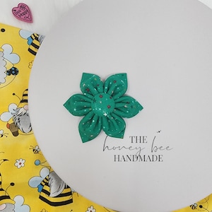 May include: A green fabric flower with white stars and red accents. The flower is handmade and has the text "THE honey bee HANDMADE" written below it.