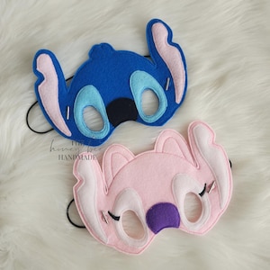 May include: Two felt character masks. One is blue with light blue and pink accents, the other is pink with white and purple accents. Both have eye holes, black elastic bands, and silver accents. The text "the honey bee handmade" is visible.
