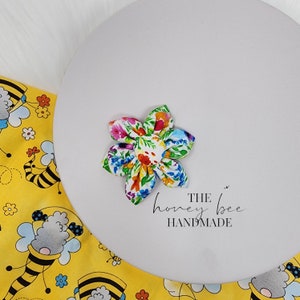 May include: A colourful floral fabric flower hair clip with a white background. The clip has a white centre and is surrounded by a variety of colours, including pink, blue, yellow, and green. The text "THE honey bee HANDMADE" is printed below the flower.