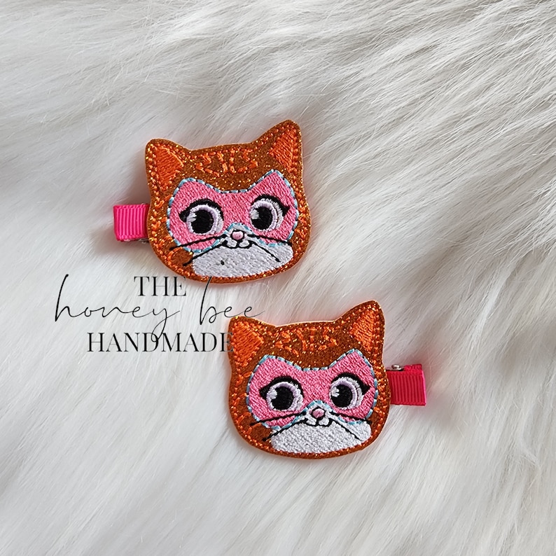 May include: Two orange glitter cat hair clips with pink and white embroidered faces. The text "THE honey bee HANDMADE" is visible in the image.