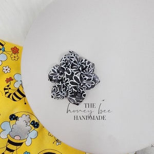May include: A black and white floral fabric flower with a white background and black floral pattern. The flower is on a white surface with the text "THE honey bee HANDMADE".