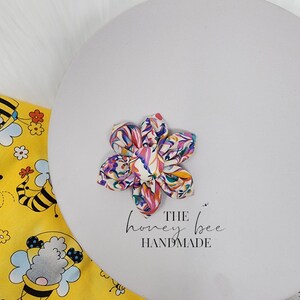 May include: A colourful floral fabric flower hair bow with a white centre. The bow is handmade and has the text "THE honey bee HANDMADE" printed below it.