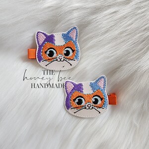 May include: Two white felt cat hair clips with orange, purple, and blue embroidered details. The cat has large eyes and a small nose. The clips are attached to orange alligator clips.