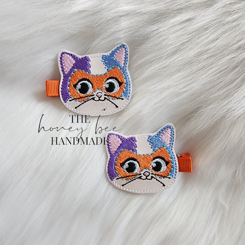May include: Two white felt cat hair clips with orange, purple, and blue embroidered details. The cat has large eyes and a small nose. The clips are attached to orange alligator clips.