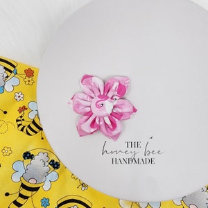 May include: Pink fabric flower with a flamingo print. The flower is handmade and has a button centre. The text "THE honey bee HANDMADE" is printed below the flower.