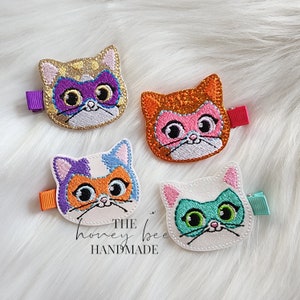 May include: Four colourful cat head hair clips with glitter and embroidered details. Each clip features a different colour scheme and a unique cat face with large eyes. The text "THE honey bee HANDMADE" is visible at the bottom of the image.