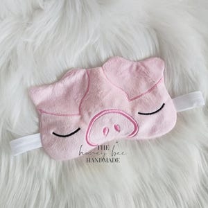 May include: Pink sleep mask with a pig face embroidered on it. The mask has a white elastic strap. The text "THE honey bee HANDMADE" is printed on the mask.