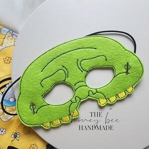 May include: Green felt mask with yellow stitching around the edges, depicting a cartoon skull with eye holes. The mask has two metal fasteners on the sides and is attached to a black elastic band. The text "THE honey bee HANDMADE" is visible below the mask.