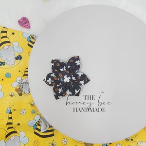 May include: A black and white fabric flower with a ghost and pumpkin pattern. The flower is handmade and has the text "THE honey bee HANDMADE" written below it.