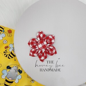 May include: Red and white gingham fabric flower with a white centre. The flower is handmade and has a simple, elegant design. The text "THE honey bee HANDMADE" is written below the flower.