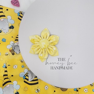 May include: A yellow fabric flower with a bee theme. The flower is on a white background with the text "THE honey bee HANDMADE".