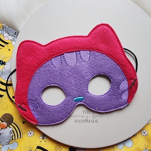 Può includere: Una maschera da gatto in feltro fatta a mano nei toni del rosa e del viola. La maschera presenta dettagli ricamati, fori per gli occhi e un piccolo naso blu. Il testo "THE honey bee HANDMADE" è visibile.