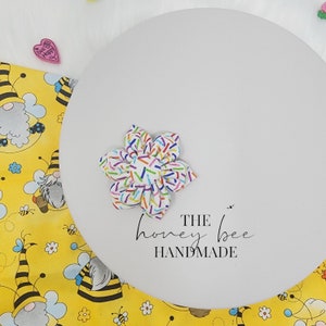 May include: A white ceramic plate with the text "THE honey bee HANDMADE" printed on it. A colourful fabric flower with white, pink, blue, green, and yellow sprinkles is on the plate.