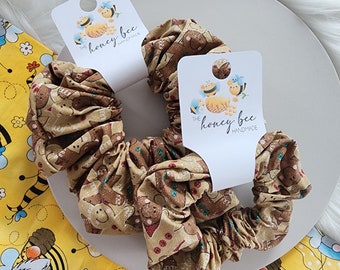 May include: Two brown scrunchies with a gingerbread man print and gold glitter. The scrunchies are on a white plate with a yellow and black fabric background. The scrunchies have tags that say "The Honey Bee Handmade".