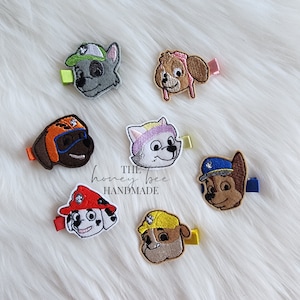May include: Set of seven embroidered felt hair clips featuring characters from the cartoon show Paw Patrol. The characters include Chase, Marshall, Skye, Rocky, Rubble, Zuma, and Everest. The clips are all different colours and have a variety of details, such as hats, goggles, and badges.