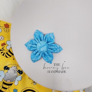 May include: A light blue fabric flower with a white pattern. The flower is handmade and has a simple, elegant design. The text "THE honey bee HANDMADE" is printed below the flower.